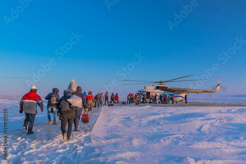 A polar aviation helicopter on a wooden heliport in the northern tundra, not far from a drilling oil well. The drillers and oil workers are dedicated and equipment is being loaded. Winter polar day. photo