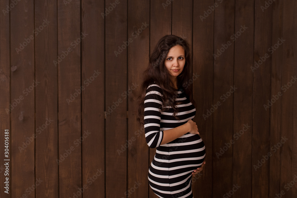 Pregnant woman in stripe dress holds hands on belly on a dark brown background. Pregnancy, maternity, preparation and expectation concept. Beautiful tender mood photo of pregnancy Pregnant woman in stripe dress holds hands on belly on a dark brown background. Pregnancy, maternity, preparation and expectation concept. Beautiful tender mood photo of pregnancy