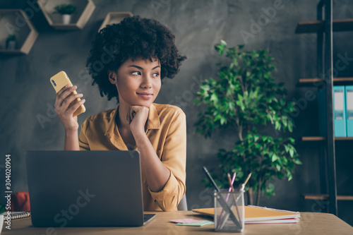 Photo of cheerful positive mixed-race dreamy worker looking wistfully into window sitting at desktop with laptop and pens on table