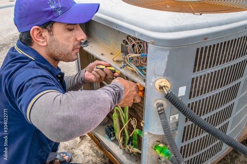 a professional electrician man is fixing the heavy unit of an air conditioner at the roof top of a building and wearing blue uniform and head cap photo