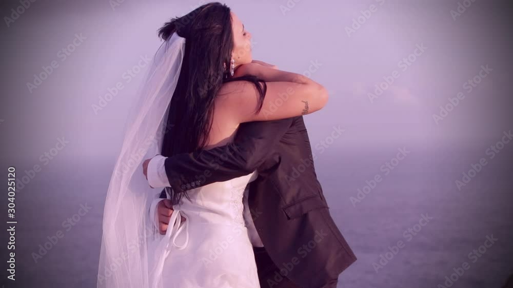 Happy married newlywed couple close up showing affection by hugging kissing touching carressing each other on wedding day at sunset beach wedding zoom magenta color grade panaromic HD wide love scene