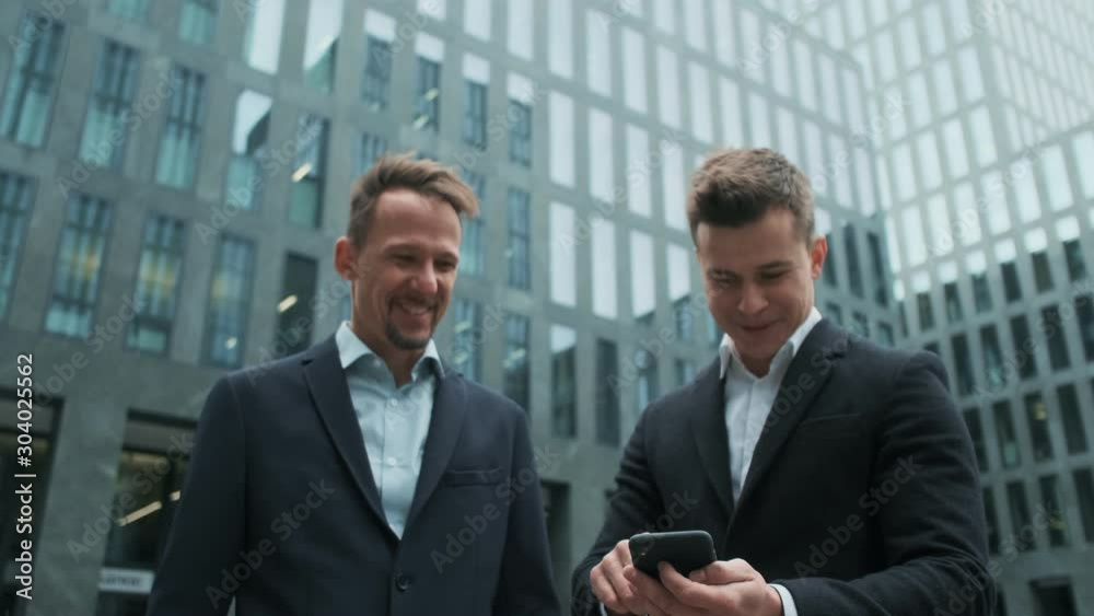 Caucasian young good looking man in business style tapping on his smartphone and showing something on it to his friend and business partner or colleague outdoor in the city center. Caucasian young good looking man in business style tapping on his smartphone and showing something on it to his friend and business partner or colleague outdoor in the city center.