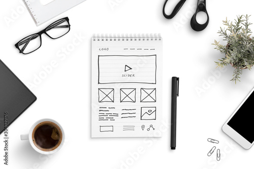 Sketch of a website on a paper writing pad, surrounded by smart phone, cup of coffee, glasess, plant. Office desk, top view, flat lay. photo