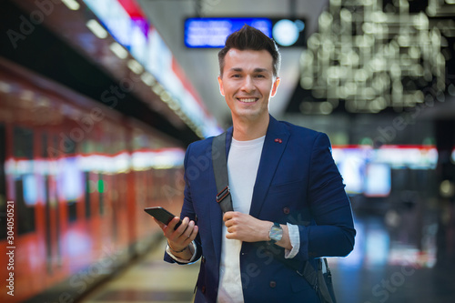 Young stylish handsome man in suit standing on metro station, holding smart phone in hand, scrolling and texting, smiling and laughing. Futuristic bright subway station. Finland