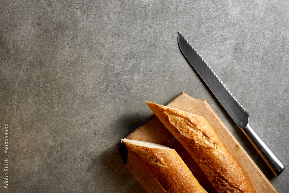 Two halves of a fresh baguette, a wooden board and a knife on a gray stone surface. Top view.