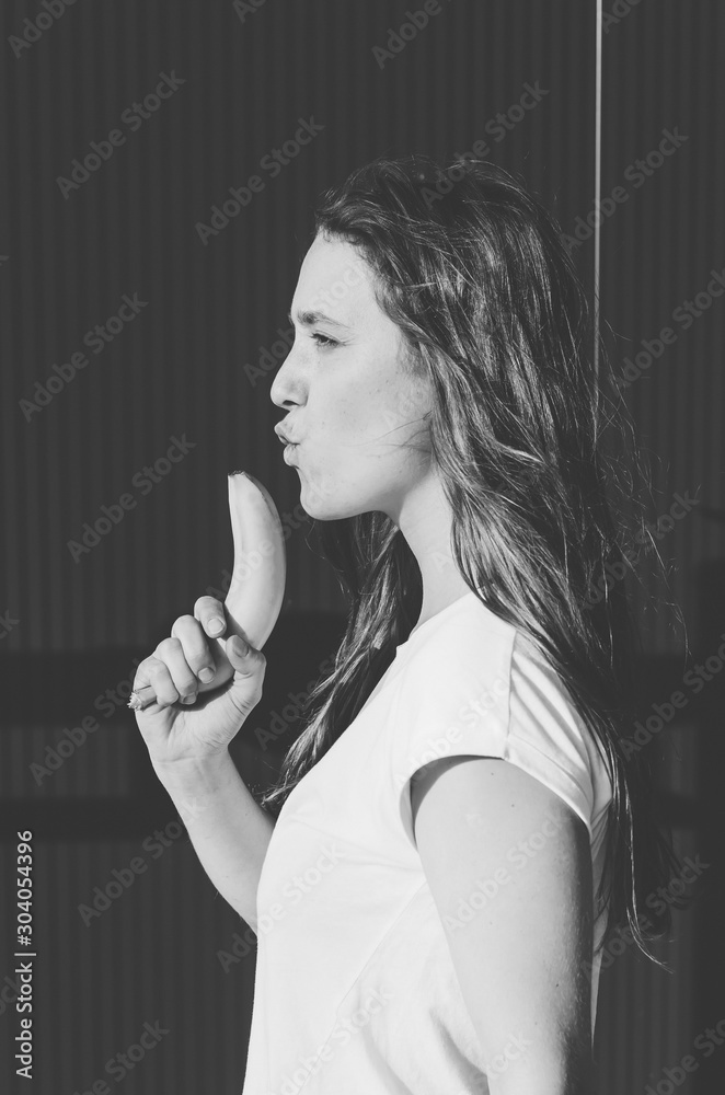 Fototapeta premium Black and white shot of Attractive young woman holding banana like gun on grey wall