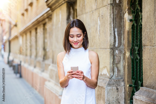 Beautiful asian woman using an application to send an sms message in her smartphone device. Happy young Asian woman with smartphone standing in the street © Graphicroyalty