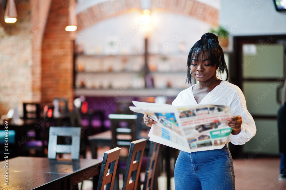 Stylish african american women in white blouse and blue jeans posed at cafe with newspaper.