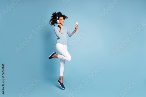 Turned full length body size photo of cheerful positive cute nice pretty girlfriend jumping up in white pants listening music isolated pastel color blue background with phone