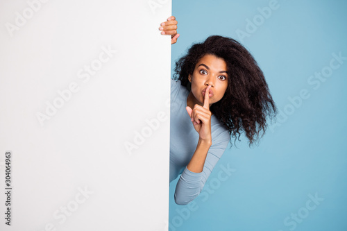 Photo of casual curly wavy beautiful brown haired girl showing you shh sign with forefinger touching her lips looking out of white banner isolated blue pastel color background