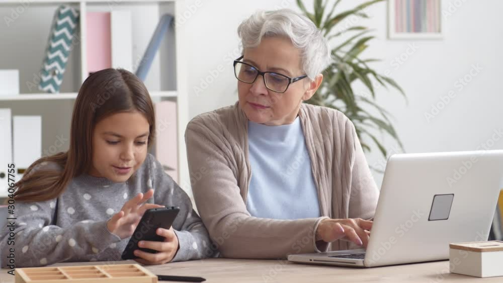 Medium shot of beautiful aged Caucasian woman with short grey hair sitting at desktop at laptop and talking to her granddaughter sitting nearby with telephone in her hands