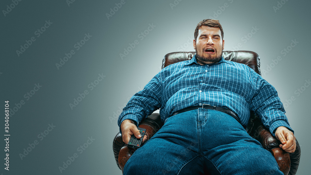 Portrait of fat caucasian man wearing jeanse and whirt sitting in a brown armchair isolated on gradient grey background. Emotional watching TV and changing channels, laughting. Overweight, carefree. Portrait of fat caucasian man wearing jeanse and whirt sitting in a brown armchair isolated on gradient grey background. Emotional watching TV and changing channels, laughting. Overweight, carefree.