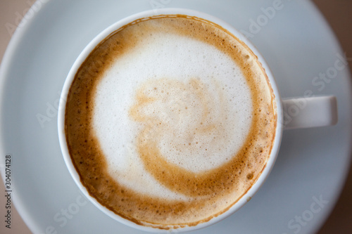 Close up white coffee cup with heart shape latte art foam on black wood table near window with light shade on tabletop at cafe.