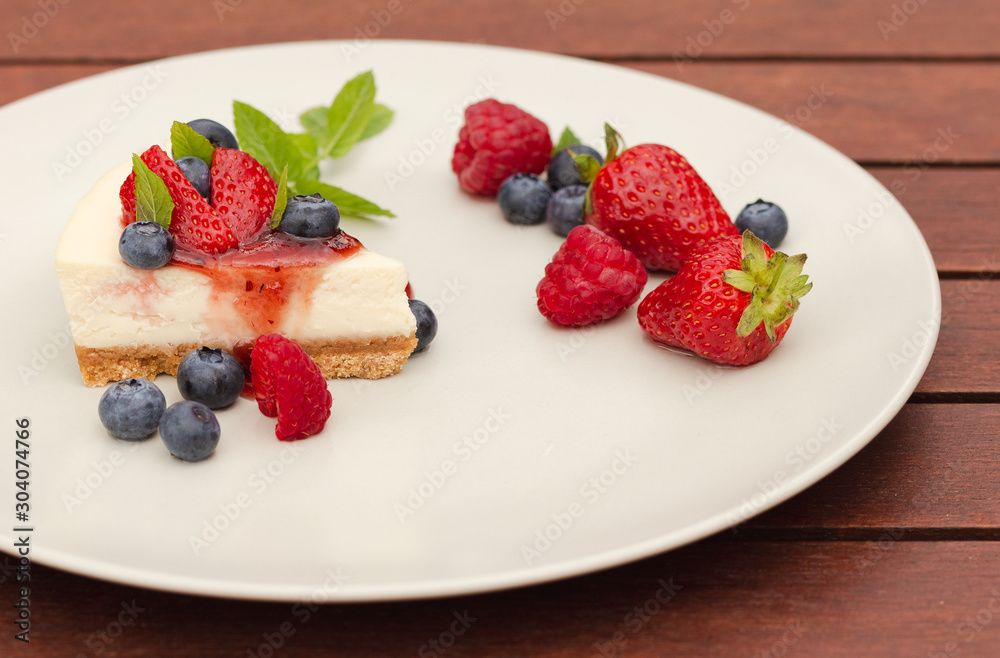 Close up of cheesecake with berries, coulis and mint on cream plate with wood background Close up of cheesecake with berries, coulis and mint on cream plate with wood background