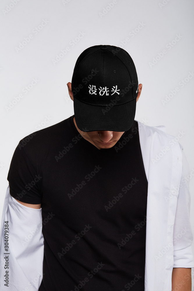 Cropped upward shot of a dark-haired man, wearing black baseball cap with  Chinese characters print, black t-shirt and white shirt. The cap peak is  closing upper part of a face. Stock Photo