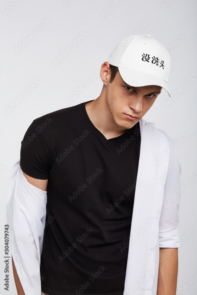 Cropped front view shot of a dark-haired man, wearing white baseball cap with Chinese characters print, black t-shirt and white shirt. He is posing on grey background and looking straight. 