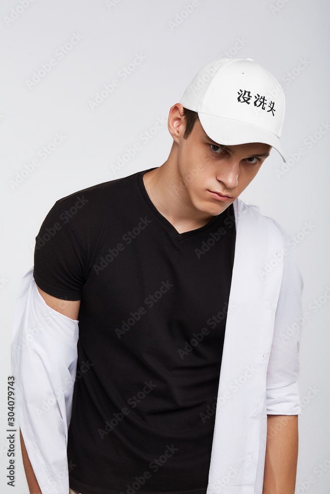 Cropped front view shot of a dark-haired man, wearing white baseball cap with Chinese characters print, black t-shirt and white shirt. He is posing on grey background and looking straight. Cropped front view shot of a dark-haired man, wearing white baseball cap with Chinese characters print, black t-shirt and white shirt. He is posing on grey background and looking straight.