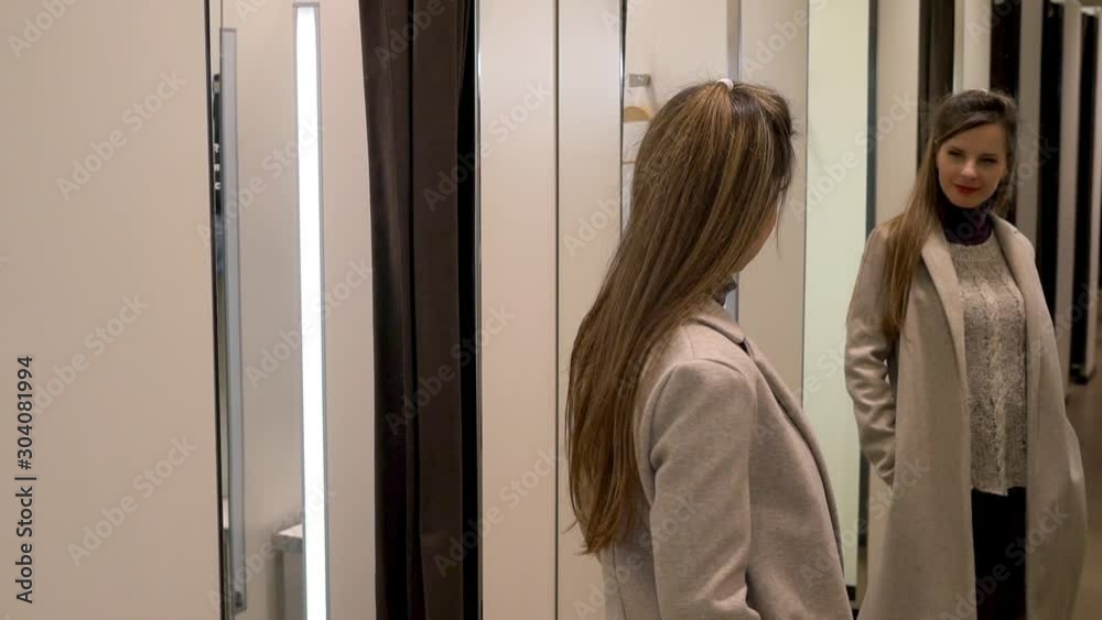 Slim shopper woman in fashion boutique shop in shopping Mall tries on modern trendy beige coat in store fitting room and looks in mirror, slow motion. Slim shopper woman in fashion boutique shop in shopping Mall tries on modern trendy beige coat in store fitting room and looks in mirror, slow motion.