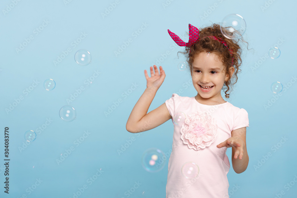 Portrait of a cheerful little girl isolated over blue background, blowing soap bubbles, having fun. Space for text. Horizontal view.