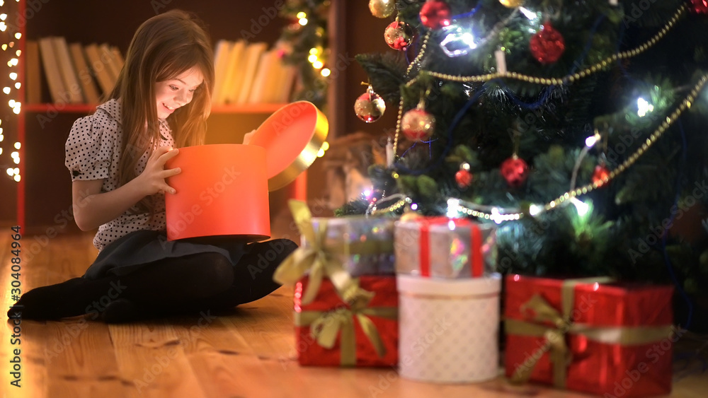 A cute girl sitting on the floor near the Christmas tree opens a box with a gift, smiles joyfully and is surprised. Christmas and New Year.