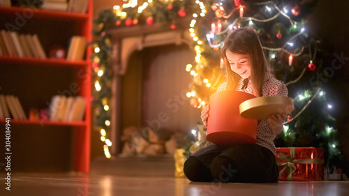 A cute girl sitting on the floor near the Christmas tree opens a box with a gift, smiles joyfully and is surprised. Christmas and New Year.