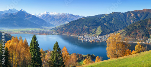 Panoramic view of beautiful autumn scene in the Alps with crystal clear Zeller lake, golden leaves and famous Kitzsteinhorn on a sunny day with blue sky in fall, Zell am See, Salzburger Land, Austria
