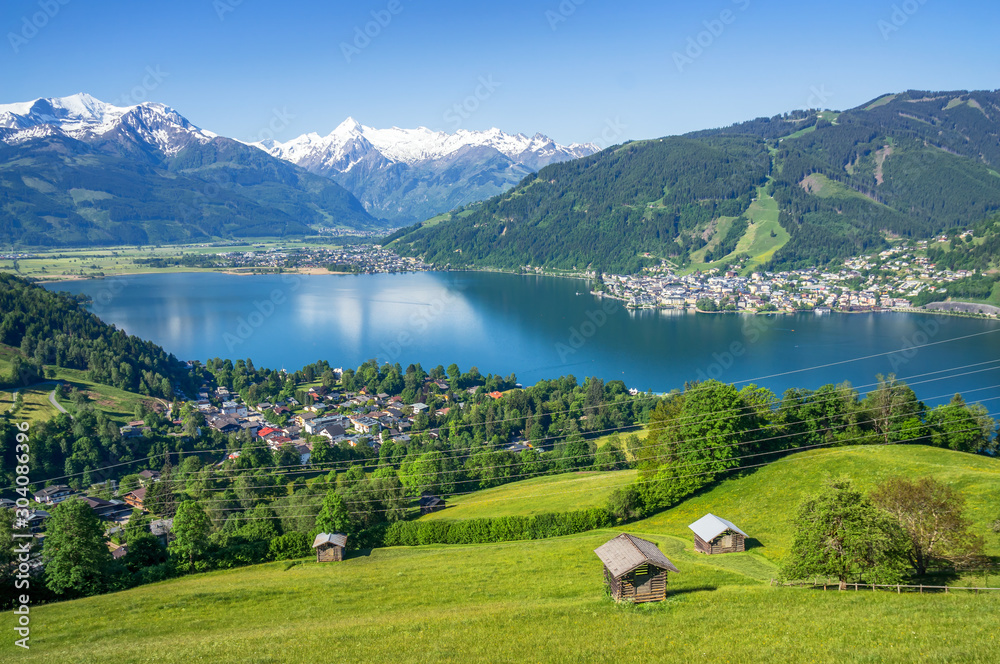 Fototapeta premium Panoramiczny widok na piękne krajobrazy Alp z czystym jeziorem, zieloną łąką, kwitnącymi kwiatami, tradycyjnymi alpejskimi domkami w słoneczny dzień z błękitnym niebem wiosną, Zell am See, Salzburger Land, Austri
