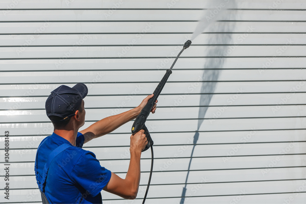 Worker in overalls washes a white wall from a siding with a water gun