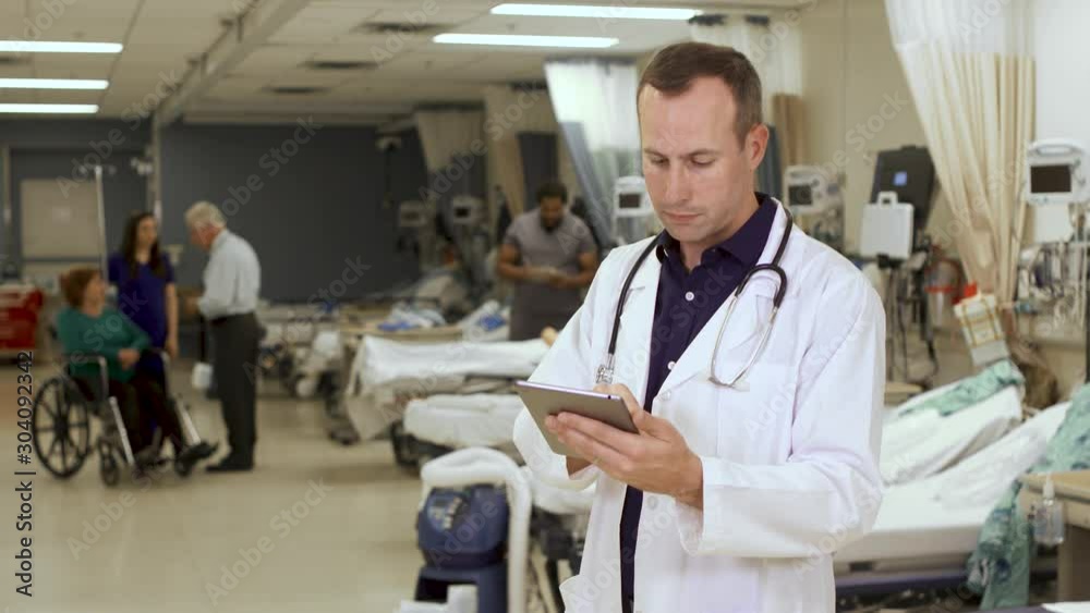 Tech savvy doctor uses a tablet computer in a hospital with nurses and patient in a wheelchair in the background. Tech savvy doctor uses a tablet computer in a hospital with nurses and patient in a wheelchair in the background.