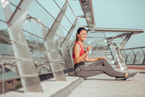 active woman sitting on the bridge after workout and drinking water from the bottle