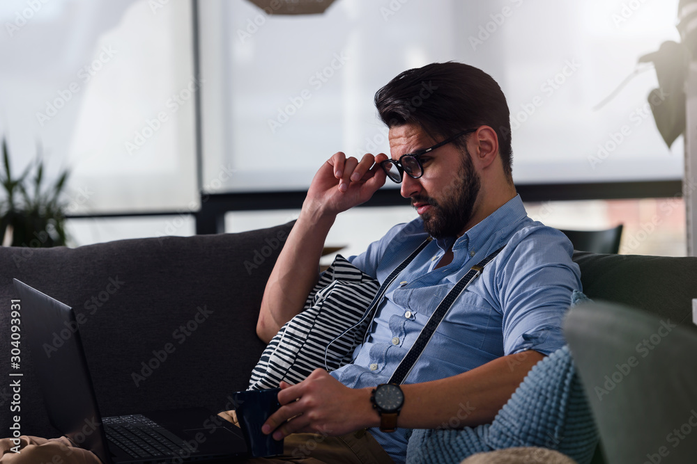 Fototapeta premium Young bearded businessman working on laptop computer while sitting on sofa at his home office