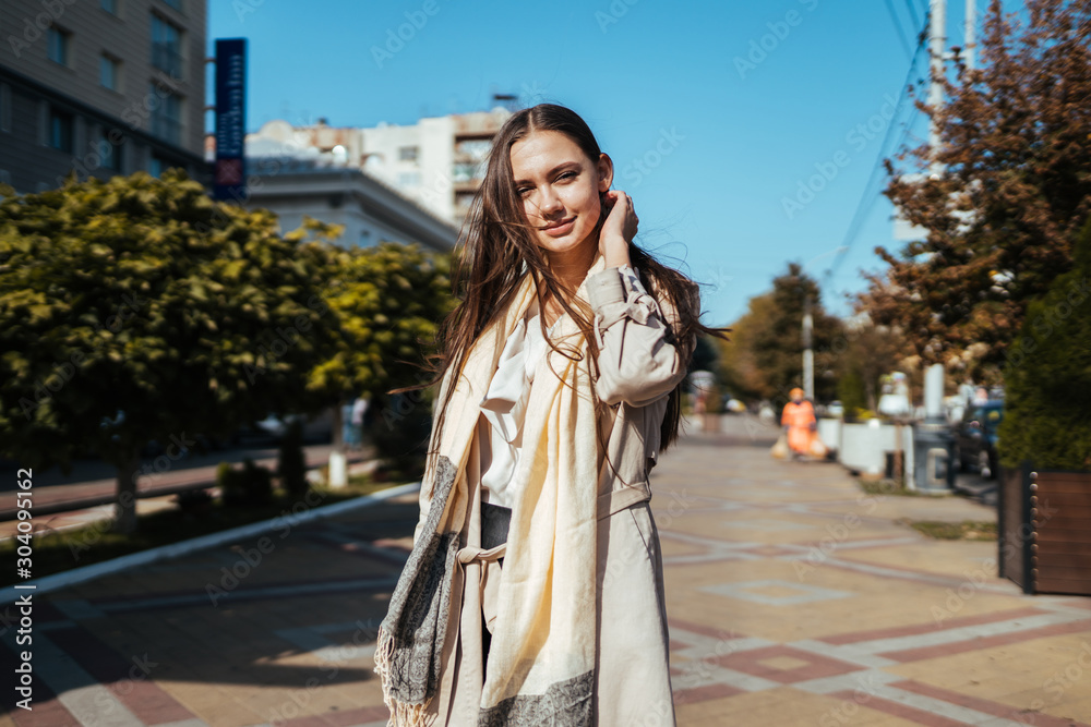 Obraz premium European woman in a cloak with long hair, straightens them by the ear on the street of a European city