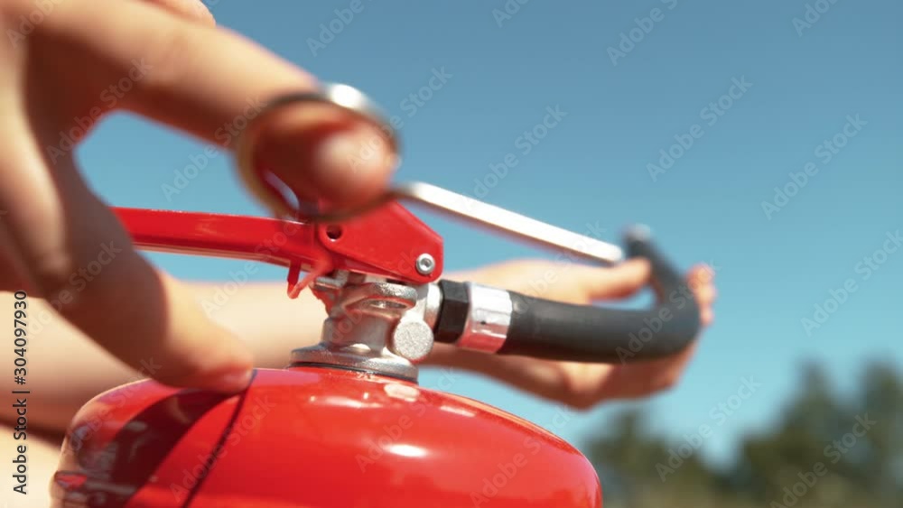 SLOW MOTION, MACRO, DOF: Young woman pulls safety pin out of a fire extinguisher before using it in an outdoor fire drill. Woman pulls out the safeguard and uses a portable fire extinguisher outdoors