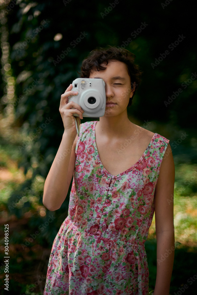Adorable preteen girl holding instant camera and taking photo closing eyes in green park