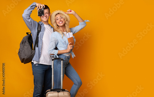 Cheerful couple with suitcase and tickets posing over yellow background