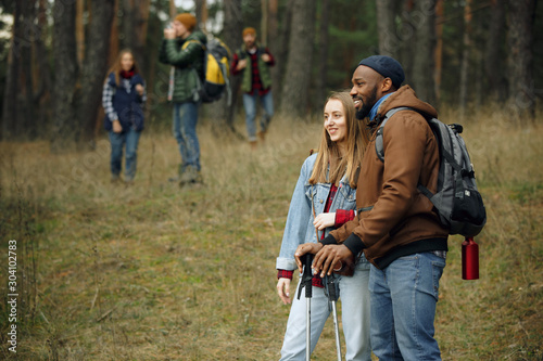 Group of friends on a camping or hiking trip in autumn day. Men and women with touristic backpacks going throught the forest, talking, laughting. Leisure activity, friendship, weekend.