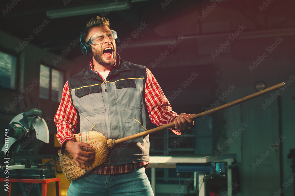  Young man on break at work has a headphones and holds a broom in his hands and pretending to play the guitar - obrazy, fototapety, plakaty 