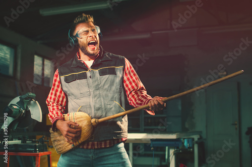  Young man on break at work has a headphones and holds a broom in his hands and pretending to play the guitar