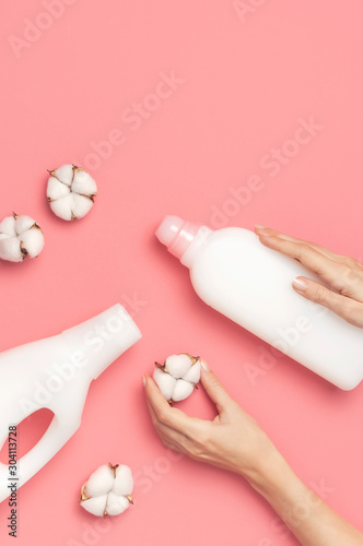 Eco cleaning concept. Women's hands hold white plastic packaging of laundry detergent, liquid powder, washing conditioner, cotton flowers on pink background. Flat lay top view. Bio organic product