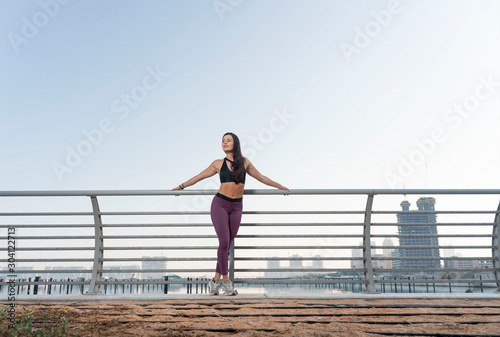 Fototapeta Naklejka Na Ścianę i Meble -  Beautiful happy brunette fitness model facing side to camera wearing a black long sleeve top and purple tights pose on a fence with a cityscape background on a bright sunny day