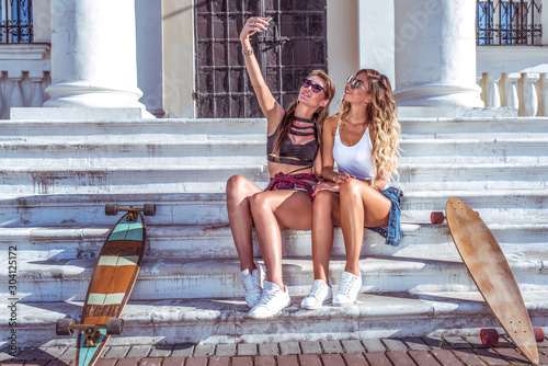 Two girls girlfriends beautiful fashionable  taking pictures phone  selfie  video call smartphone  happy smiling  resting recording video message. Social network app Internet. Longboard summer city.