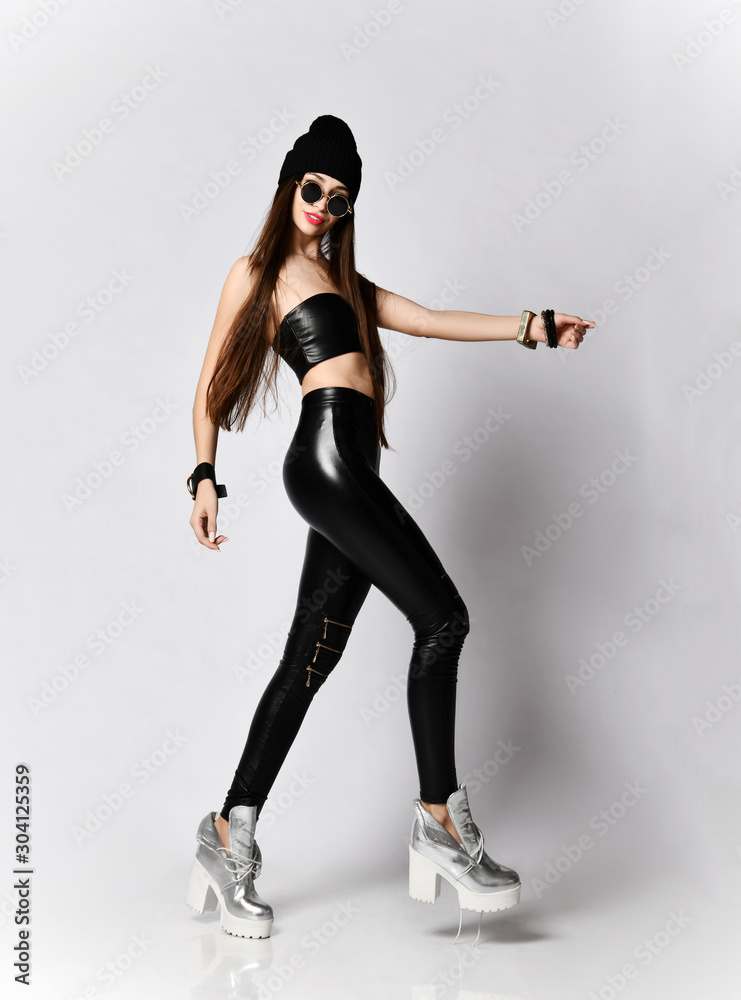 Young hipster woman in black hat, tight leather leggings and round sunglasses is walking, passing by, looking at us Young hipster woman in black hat, tight leather leggings and round sunglasses is walking, passing by, looking at us