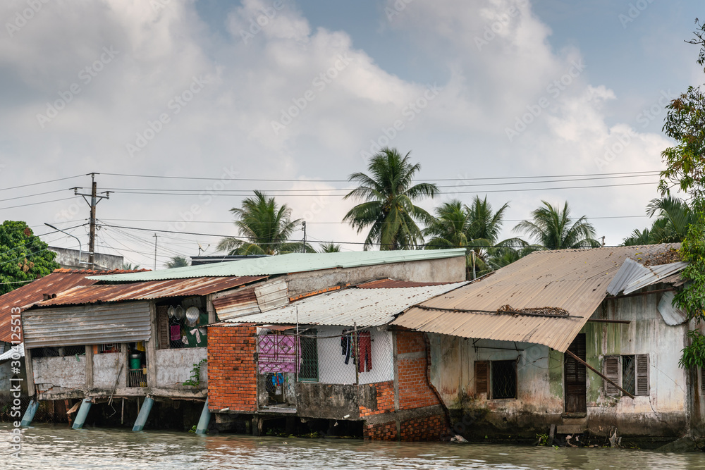 Cai Be, Mekong Delta, Vietnam - March 13, 2019: Along Kinh 28 canal. Row of slum-like poor houses built on stilts and on shore with corrugated roof plates under blue cloudscape and green foliage.
