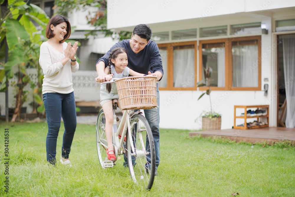 Young Asian family love, father and mother teach and training daughter to ride a bike on grass field at outdoor of home. girl train to ride a bike by her parent which smiling and felling happy Young Asian family love, father and mother teach and training daughter to ride a bike on grass field at outdoor of home. girl train to ride a bike by her parent which smiling and felling happy