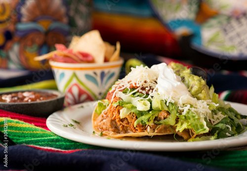 Crispy tortilla tostada with chicken, beans, lettuce, cheese, guacamole, sour cream, and salsa. Served with chips and dipping salsa styled on a sarape. photo