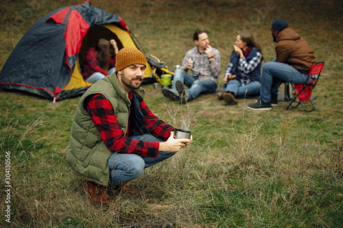Group of friends on a camping or hiking trip in autumn day. Men and women with touristic bag having break in the forest, talking, laughting. Leisure activity, friendship, weekend. Drink in thermos.