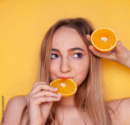 a beautiful blonde girl looks to the right, holds an orange slice in her mouth and half an orange in her hand close on a bright yellow background