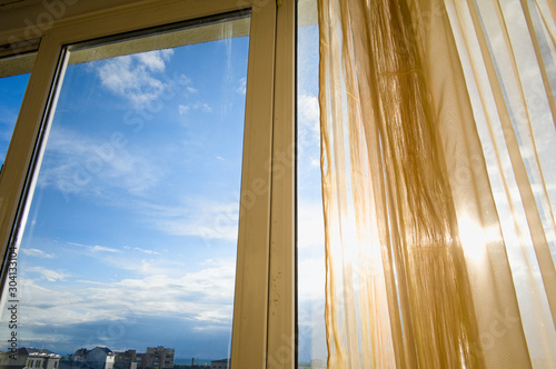 Sun shining through curtains and window. Sunlit apartment on sunset. City view and blue sky. © Vitalii Karas