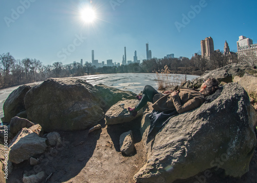 Young woman lying on rocks by frozen lake in Central Park with a view of Manhattan