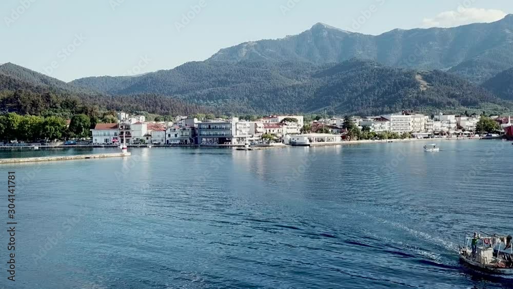 Static aerial shot of a small fishing boat passing through the frame with the town of Limenas in the background, on Thasos island, Greece.